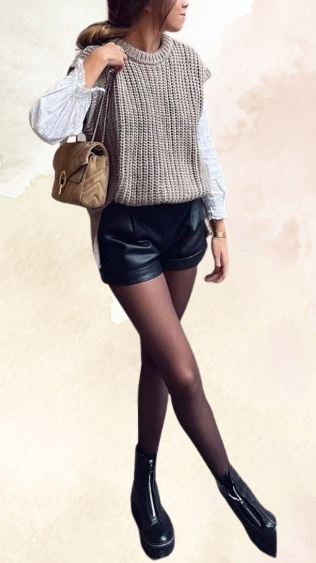 This outfit features a relaxed-fit, chunky knit sweater in a soft taupe color layered over a white, long-sleeved blouse with ruffled cuffs. Paired with high-waisted black faux leather shorts, the look is completed with sheer black tights and ankle boots for a chic, casual fall style. The overall ensemble combines textures and layers while offering a modern silhouette.