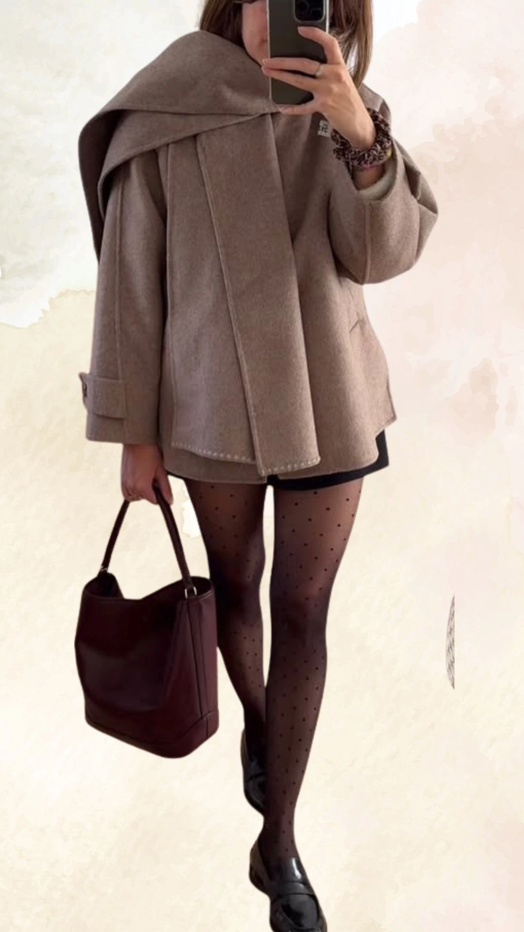 The outfit features an oversized, beige wool coat with a dramatic winged collar and folded sleeves, paired with a fitted black mini skirt. Underneath, sheer black tights with small polka dots add texture, while shiny black loafers complete the look. A dark burgundy handbag contrasts beautifully with the overall neutral palette, making this a chic casual fall ensemble.