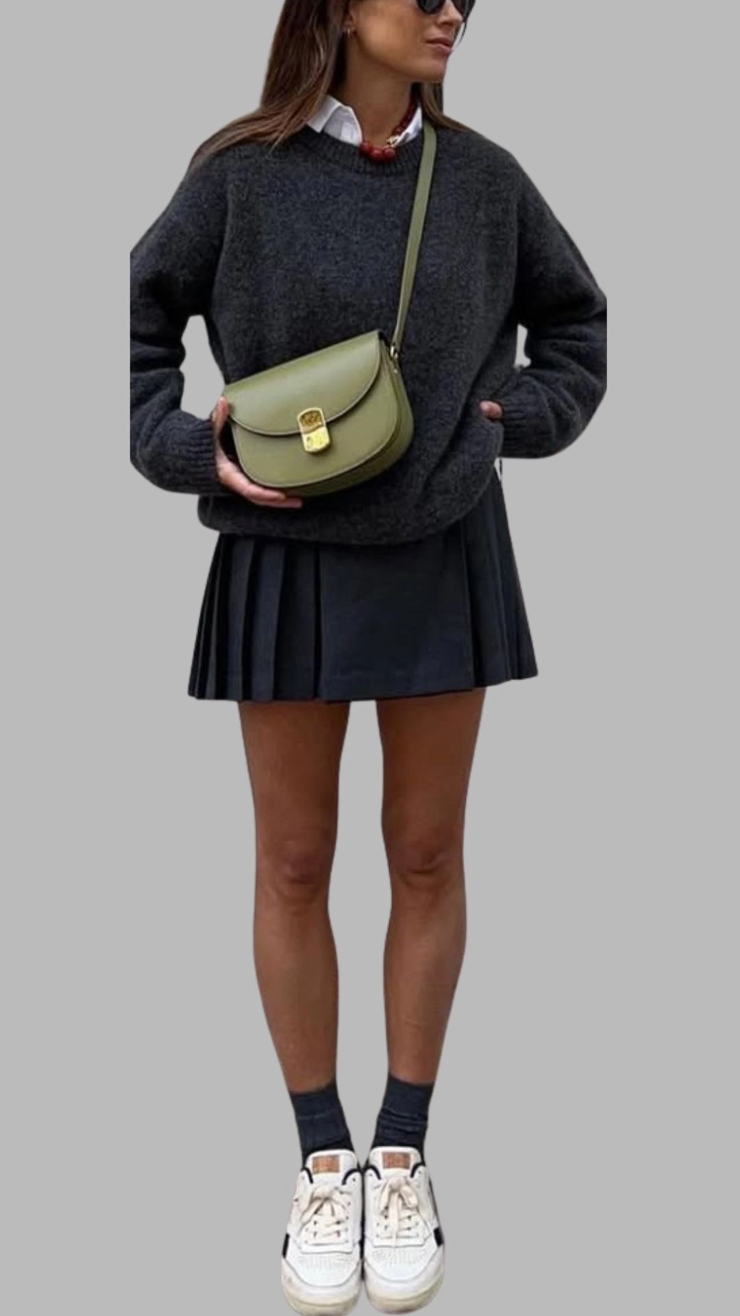 The outfit features a dark grey oversized sweater with a round neckline and long sleeves, paired with a black pleated skirt that falls above the mid-thigh. Complementing the look, the individual wears white sneakers with black accents and dark grey ankle socks, accessorized with an olive green crossbody bag. The overall ensemble balances comfort and style, showcasing a modern, casual aesthetic.