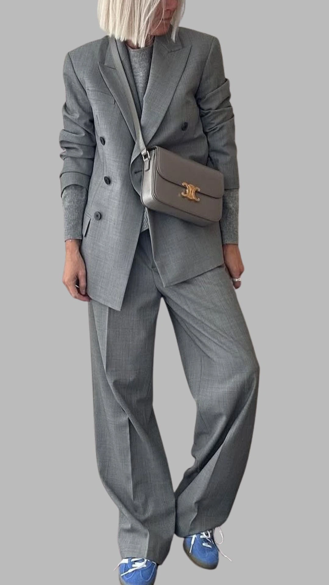 A tailored grey double-breasted blazer features structured shoulders and peak lapels, paired with matching wide-leg trousers for a polished look. Underneath, a grey knitted sweater adds texture, while a light grey crossbody bag with a gold accent completes the outfit. The ensemble is styled with blue sneakers, creating a modern blend of chic and casual aesthetics.