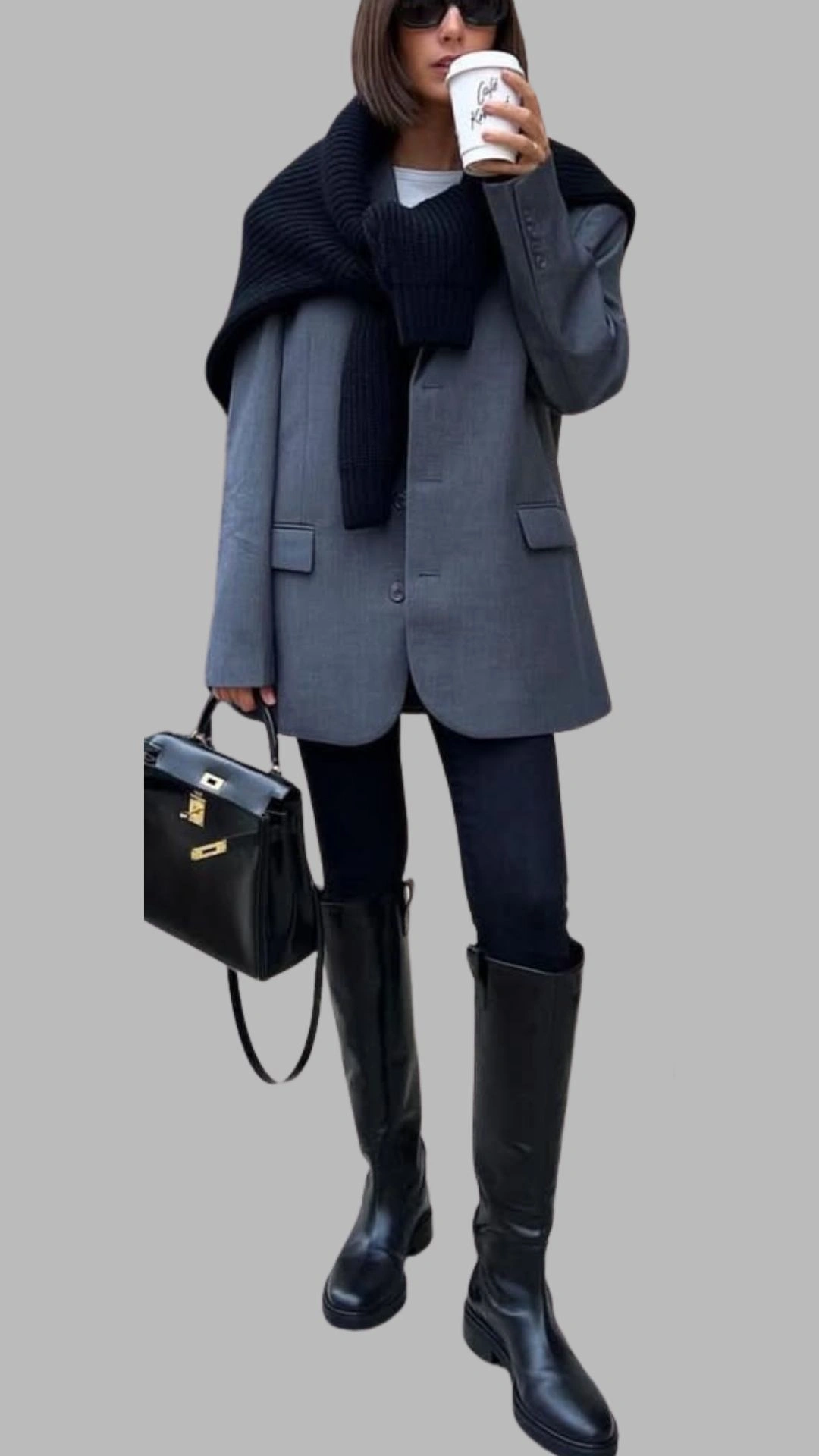 The outfit features a structured grey blazer with a single button closure and front pockets, layered over a fitted black turtleneck. Complemented by sleek black leggings and knee-high black boots, the look is accessorized with a chunky black knit scarf draped around the shoulders and a chic black handbag. This monochromatic palette emphasizes a modern, sophisticated style while ensuring comfort and versatility.