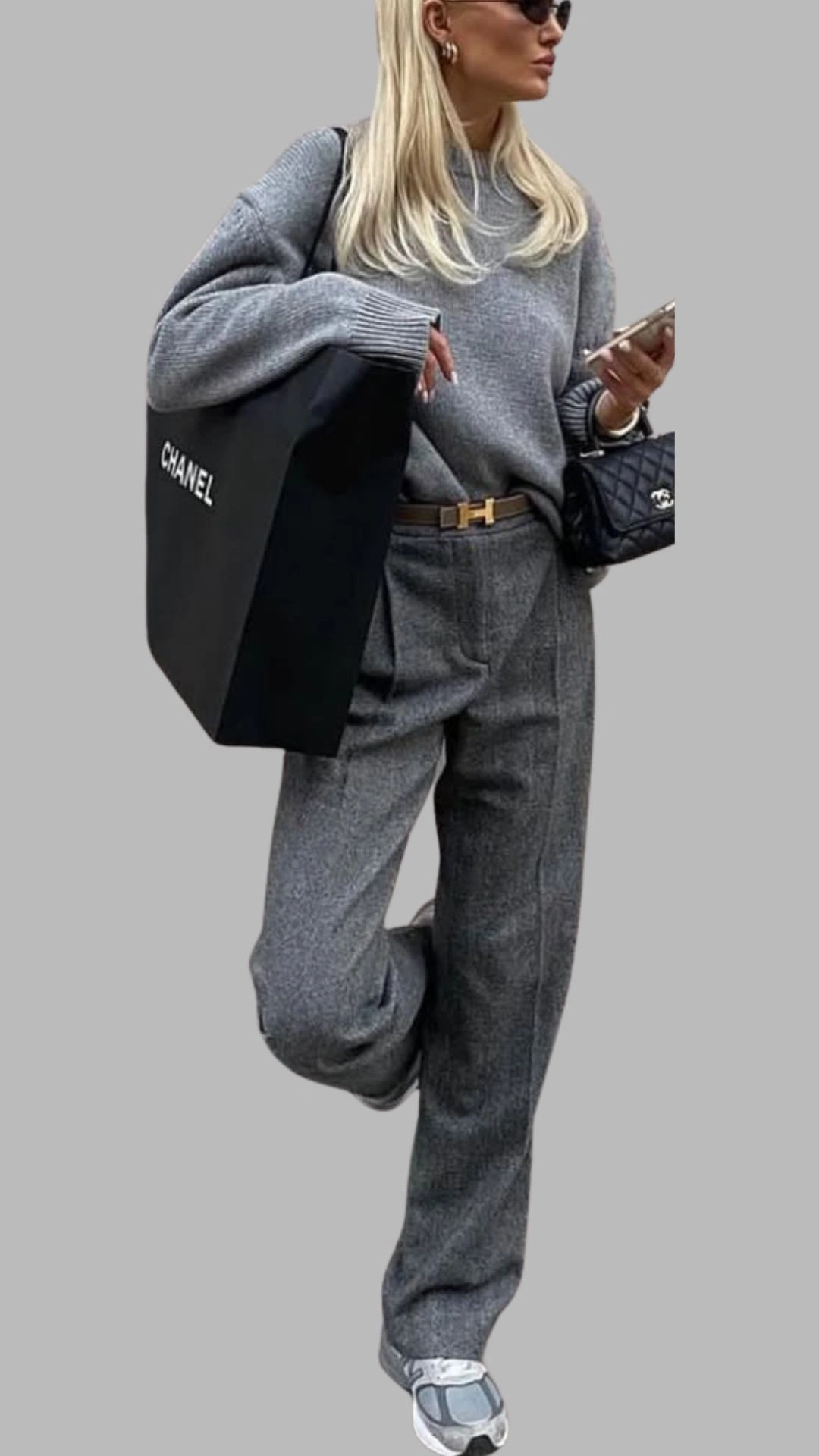 The outfit features a relaxed-fit, light grey sweater paired with high-waisted, loose-fitting grey trousers, both made from textured fabric. The look is accentuated with a brown belt and completed with chunky grey sneakers, while a large black bag adds a chic contrast. The overall palette consists of various shades of grey, creating a sophisticated yet casual ensemble.