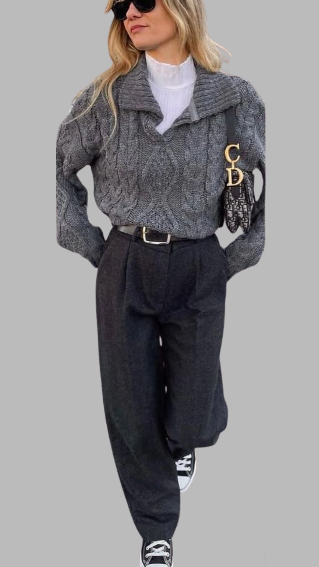 The outfit features a chunky, dark grey cable-knit sweater with a wide, folded collar, paired with high-waisted, loose-fitting charcoal trousers that taper at the hem. The look is complemented by black and white sneakers, adding a casual touch, while a stylish crossbody bag enhances the overall aesthetic.