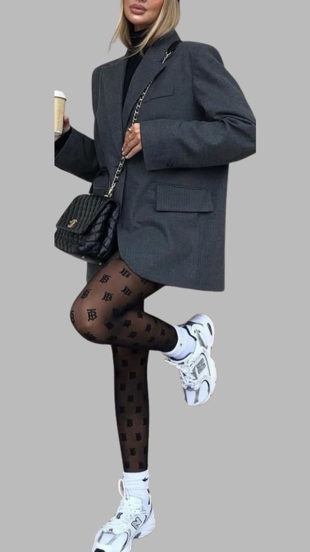 The outfit features an oversized grey blazer with a slightly structured shoulder and large front pockets, layered over a fitted black turtleneck bodysuit. Paired with patterned black tights that showcase a subtle logo design, the look is completed with chunky white sneakers for a modern, casual flair. The combination of dark and light colors creates a stylish contrast while maintaining an urban aesthetic.