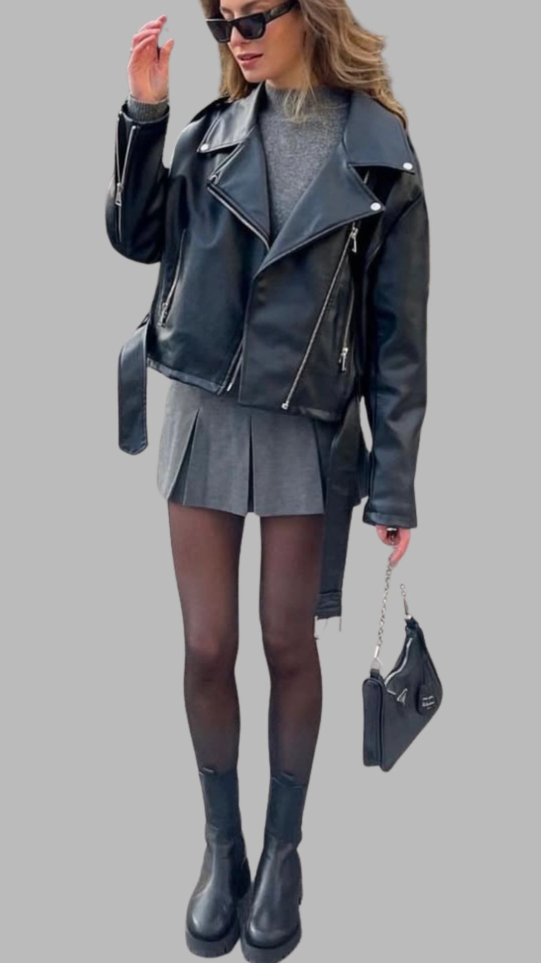 The outfit features a layered look with a fitted grey sweater peeking out from under a cropped black leather biker jacket, which has an asymmetric zipper and wide lapels. The dress underneath is a pleated, short grey skirt that adds a dynamic feel, complemented by sheer black tights and chunky black ankle boots for a stylish edge. The overall palette is monochromatic, with varying shades of grey and black, creating a sleek and modern aesthetic.