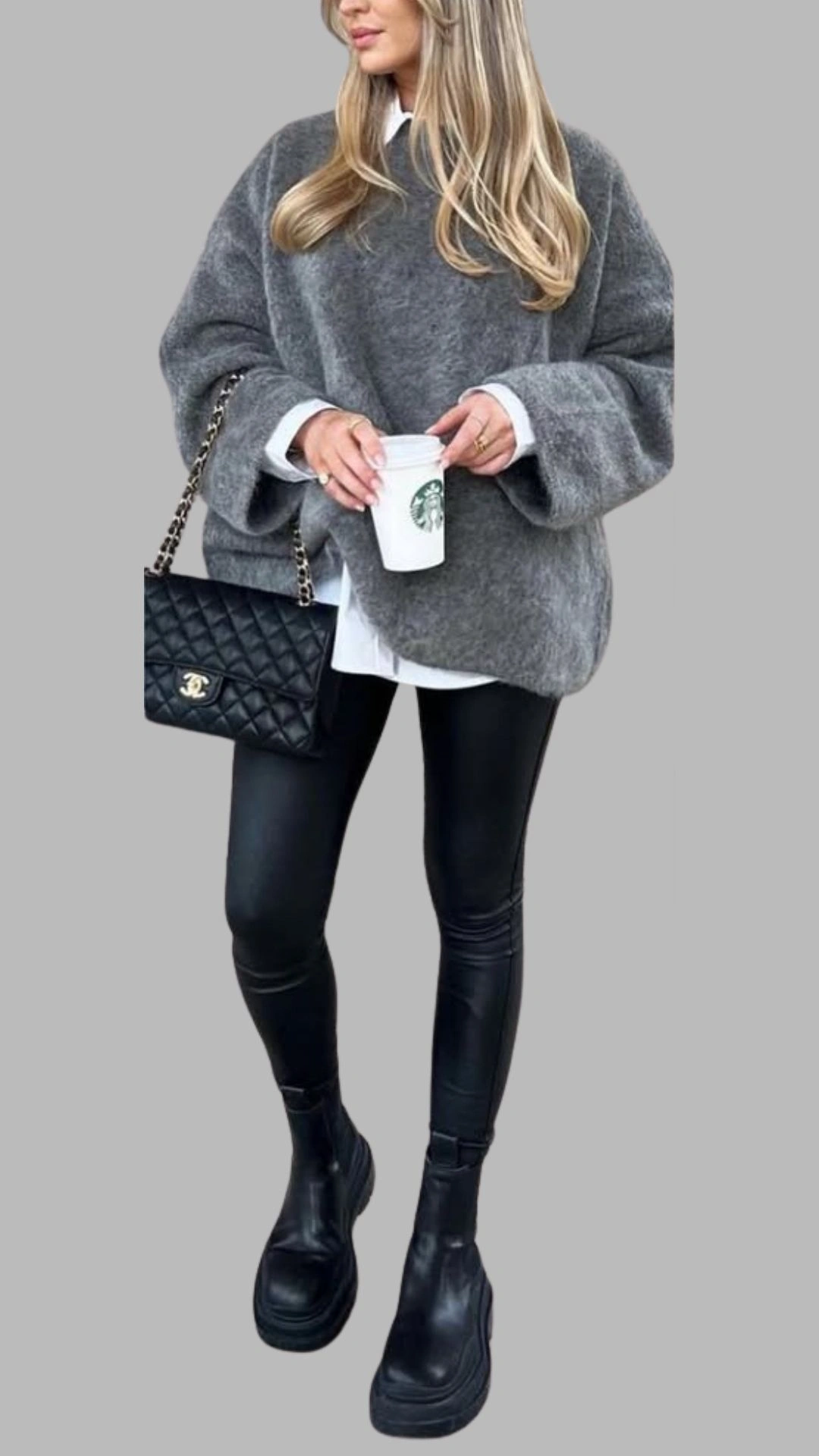 The outfit features a cozy, oversized grey sweater with a round neckline and wide, dropped sleeves, layered over a crisp white button-up shirt for a chic contrast. The look is paired with high-waisted black faux leather leggings that offer a sleek silhouette, finished with chunky black ankle boots for a bold touch. This ensemble combines texture and dimension, perfect for a modern, stylish aesthetic.