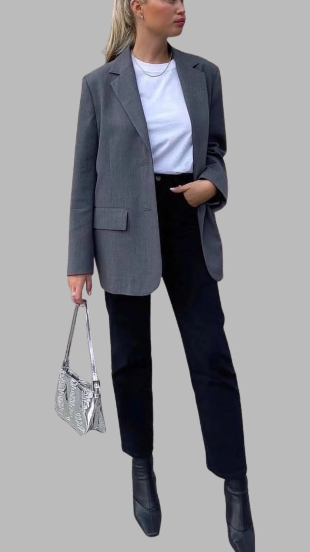 The outfit features a tailored grey blazer with a structured fit, slightly oversized shoulders, and a single front pocket, layered over a classic white t-shirt. The ensemble is paired with high-waisted, dark black trousers that taper at the ankle, complemented by pointed black ankle boots. A metallic silver handbag adds a chic touch, enhancing the modern, monochromatic palette of grey and black.