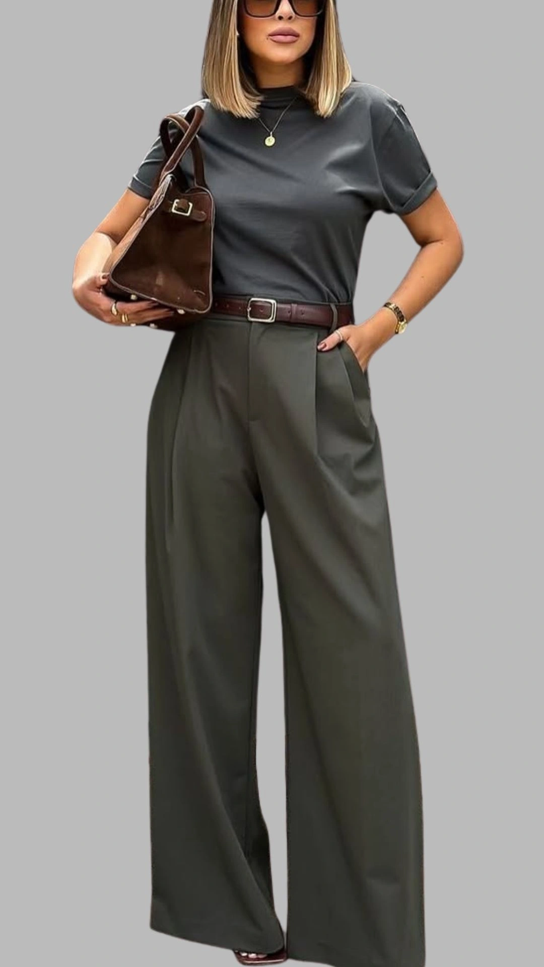 The outfit features a fitted dark grey t-shirt with a slight mock neckline and short sleeves, paired with high-waisted, wide-leg trousers in a muted olive green shade. The trousers include front pleats and practical side pockets, accentuated by a brown leather belt that defines the waist. The look is completed with a stylish brown handbag, offering a sophisticated monochromatic palette.
