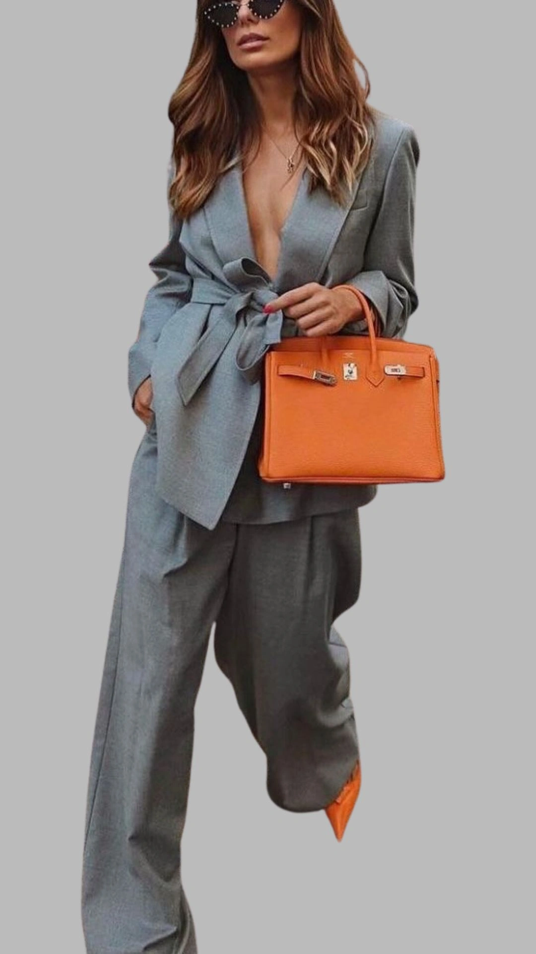 The outfit features a tailored grey blazer with a deep V-neckline and long sleeves, cinched at the waist with a matching tie, paired with loose-fit trousers that fall elegantly to the ankles. The ensemble is complemented by an orange handbag, adding a vibrant contrast, while the whole look embodies a chic, modern aesthetic. The soft texture of the grey fabric enhances the sophisticated appearance, making it suitable for both professional and stylish casual settings.