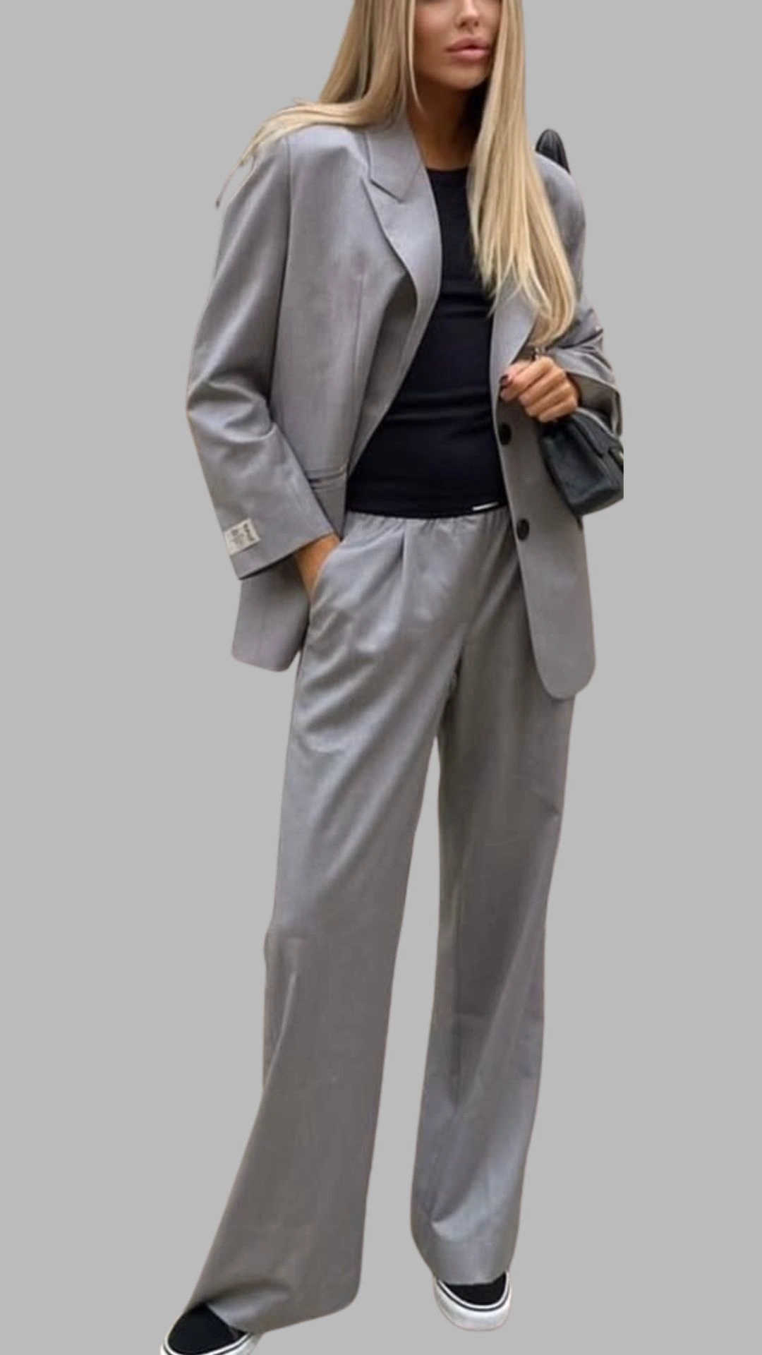 The outfit features a tailored light grey oversized blazer with structured shoulders, complemented by high-waisted, wide-leg grey trousers that drape elegantly. Underneath, a simple black fitted top provides a sleek contrast, while the addition of casual black sneakers adds a relaxed touch to the sophisticated ensemble. The overall look combines modern tailoring with comfortable elements, making it suitable for a variety of occasions.