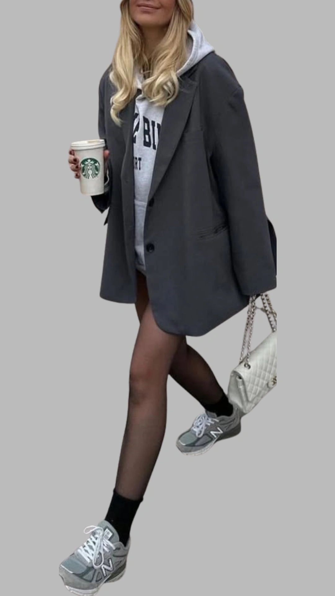 The outfit features an oversized charcoal grey blazer paired with a light grey hoodie, showcasing a relaxed yet stylish look. The blazer has a notched lapel and front pockets, while the sheer black tights and grey sneakers with white accents add a modern touch. A cream quilted handbag completes the ensemble, creating a balanced and trendy aesthetic.