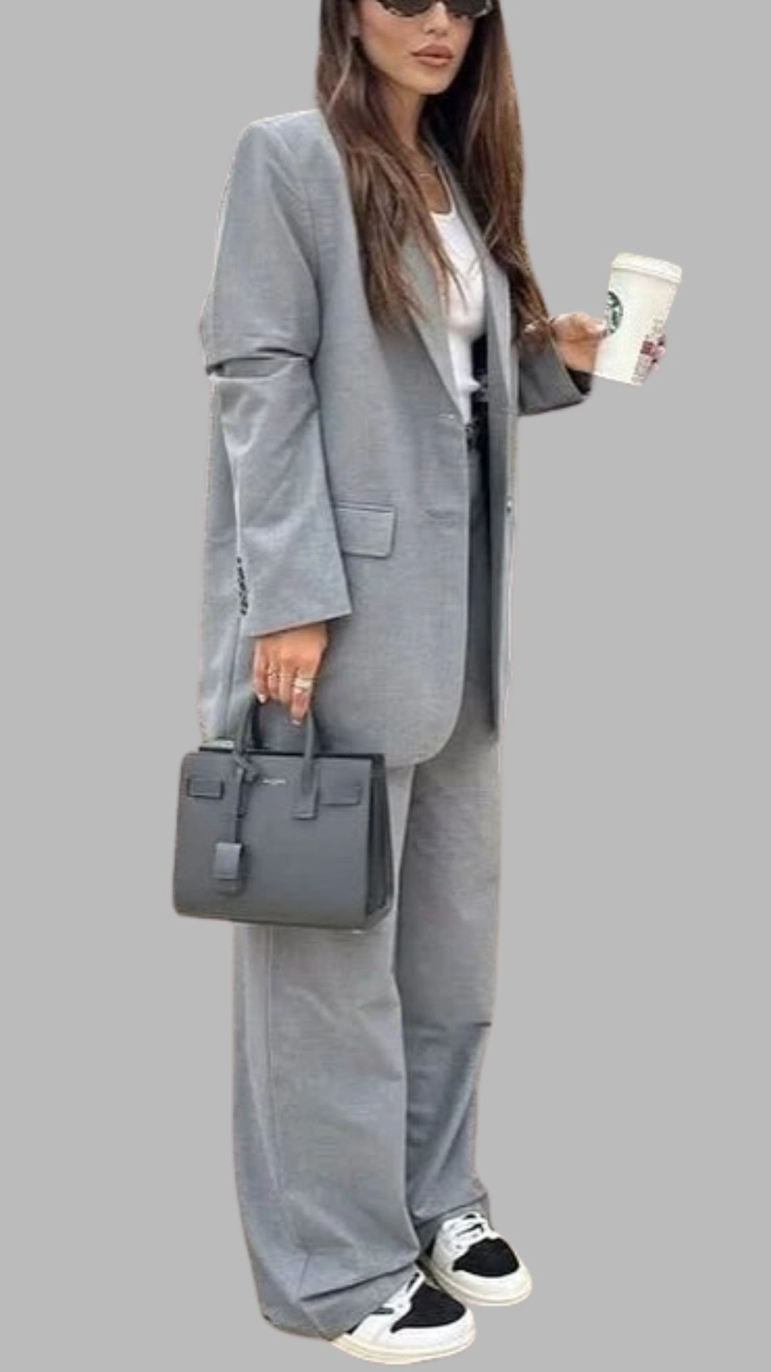 The outfit features a tailored grey oversized blazer paired with matching wide-leg trousers, creating a chic and modern silhouette. Underneath, a simple white tee adds contrast and balance, while the look is accessorized with a sleek black handbag and casual black and white sneakers, enhancing its versatility. The overall ensemble combines neutral tones with a polished yet relaxed vibe, perfect for various occasions.
