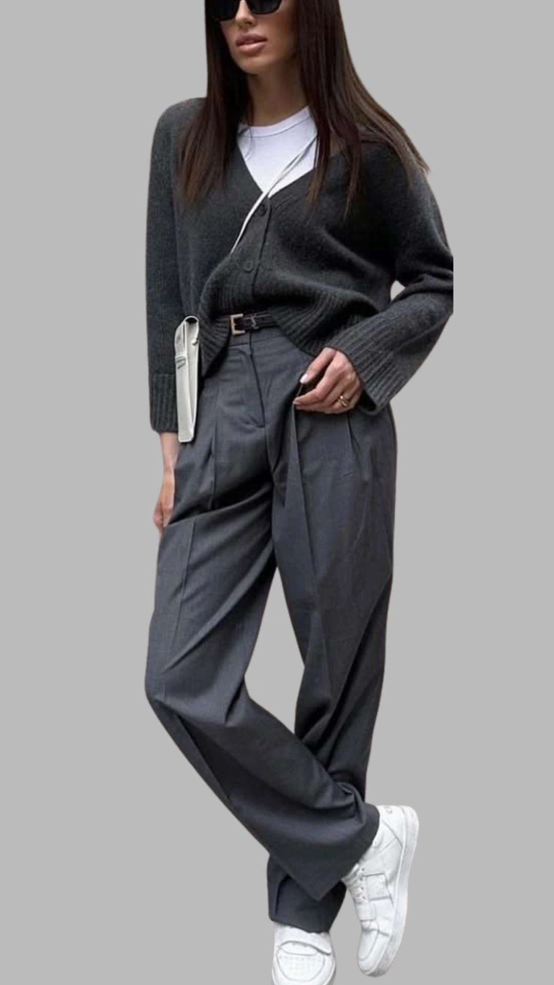 The outfit features a dark grey oversized cardigan with a V-neckline and ribbed detailing, paired with high-waisted, tailored grey trousers that exhibit a subtle pinstripe pattern. The look is completed with white sneakers, adding a casual touch, while a slim belt cinches the waist for definition. This ensemble combines shades of grey with a comfortable, stylish aesthetic.