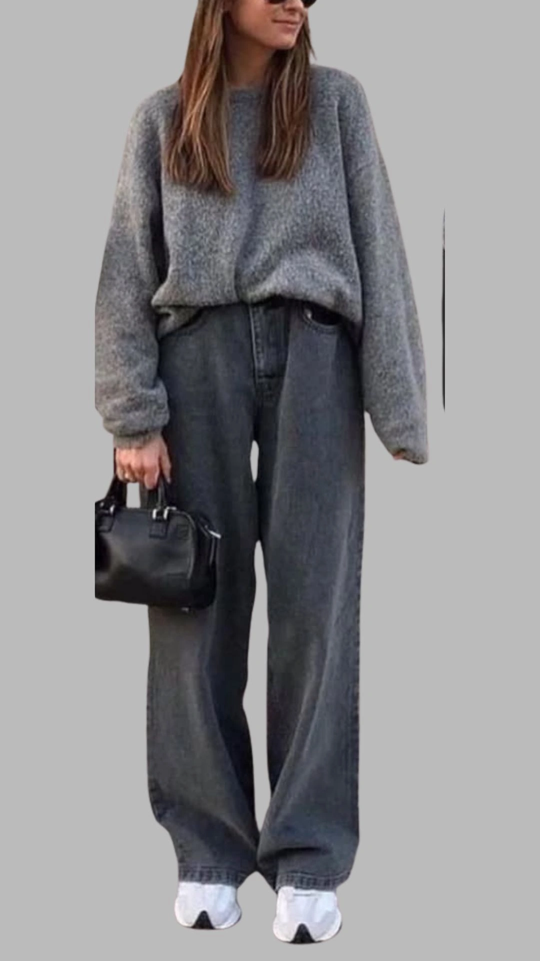The outfit features a cozy, oversized, light grey sweater with a round neckline and long sleeves, paired with high-waisted, wide-leg dark grey jeans that have a relaxed fit. The ensemble is completed with white sneakers and a small black handbag, creating a monochromatic yet stylish look with subtle contrasts in grey shades. This casual outfit is both comfortable and chic, making it versatile for various occasions.