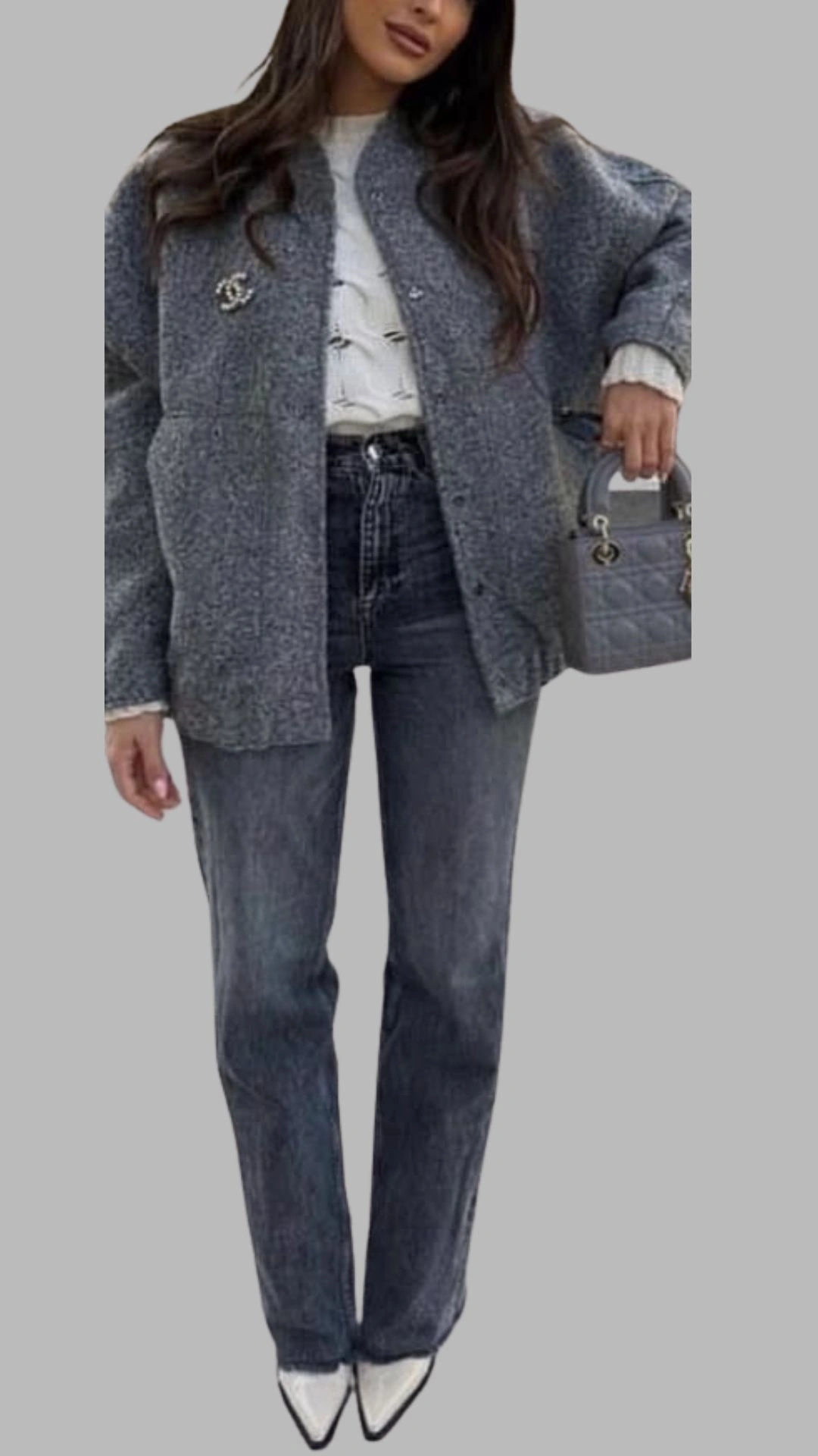 The outfit features a relaxed grey oversized jacket with a subtle texture and a statement pearl brooch on the left side, layered over a white knitted sweater. The ensemble is paired with high-waisted, dark grey straight-leg jeans, creating a sleek silhouette. Completing the look are sharp white pointed-toe heels and a small grey quilted handbag, adding a chic contrast to the neutral palette.
