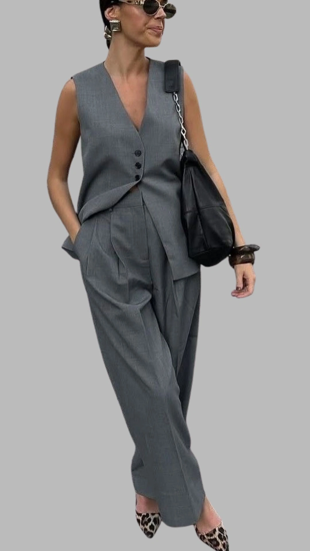 The outfit features a tailored grey vest with a deep V-neckline and three front buttons, paired with high-waisted, loose-fitting trousers that have pleated details. The ensemble combines a textured, pinstripe pattern with a sleek silhouette, complemented by stylish pointed-toe heels in a leopard print. A black handbag completes the look, enhancing its modern sophistication.