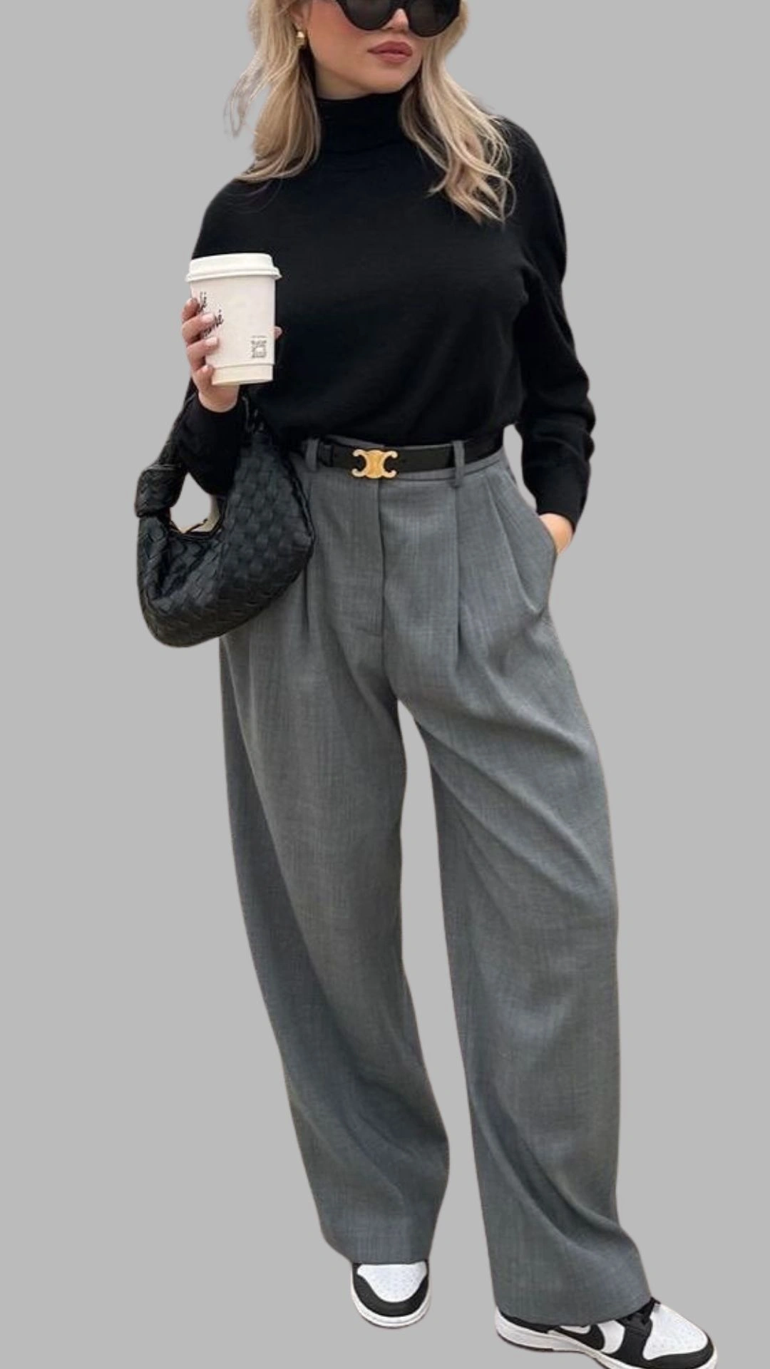 The outfit features a fitted black turtleneck sweater paired with high-waisted, light grey wide-leg trousers that have a subtle plaid pattern. The trousers are styled with a statement black belt featuring a gold buckle for added flair, complemented by a sleek black handbag and casual white sneakers with black accents.