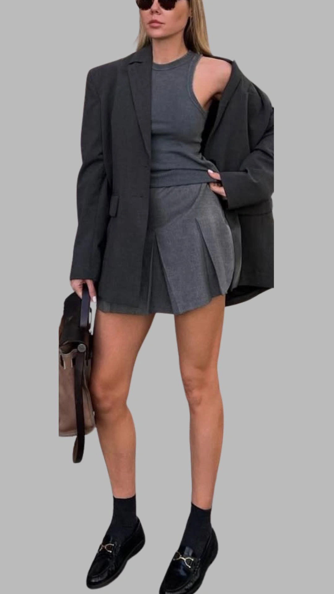 The outfit features a fitted grey tank top layered under an oversized charcoal grey blazer, adding a contemporary touch. The mini skirt showcases a pleated design in a slightly lighter shade of grey, enhancing the structured silhouette. Paired with black loafers and ankle socks, this ensemble balances chic with casual elegance.