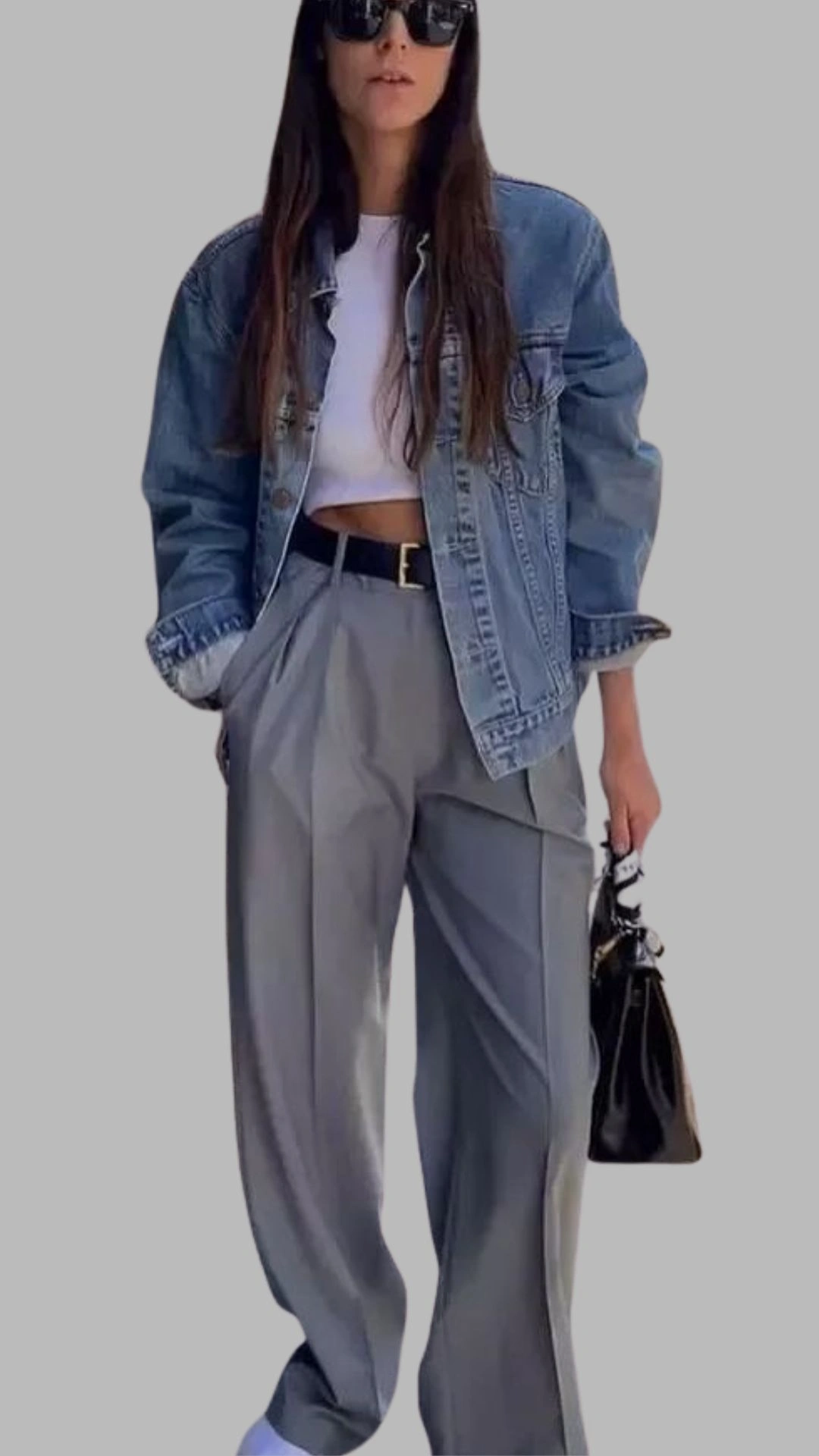 The outfit features high-waisted grey trousers with a relaxed fit and sharp pleats, offering a sophisticated look. Paired with a cropped white top and an oversized denim jacket, the ensemble balances casual and chic elements. Accessories include a black belt and a stylish black handbag, enhancing the overall aesthetic.