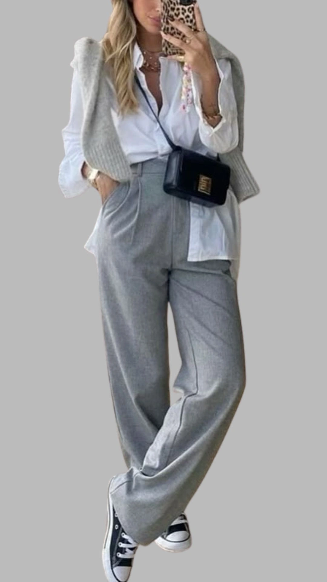The outfit features wide-legged, light grey trousers with a high waist and a relaxed fit, paired with an off-white collared shirt that peeks out at the bottom. Layered on top is a soft, light grey knitted vest, creating a sophisticated yet casual look. The ensemble is completed with classic black and white sneakers for a modern touch.
