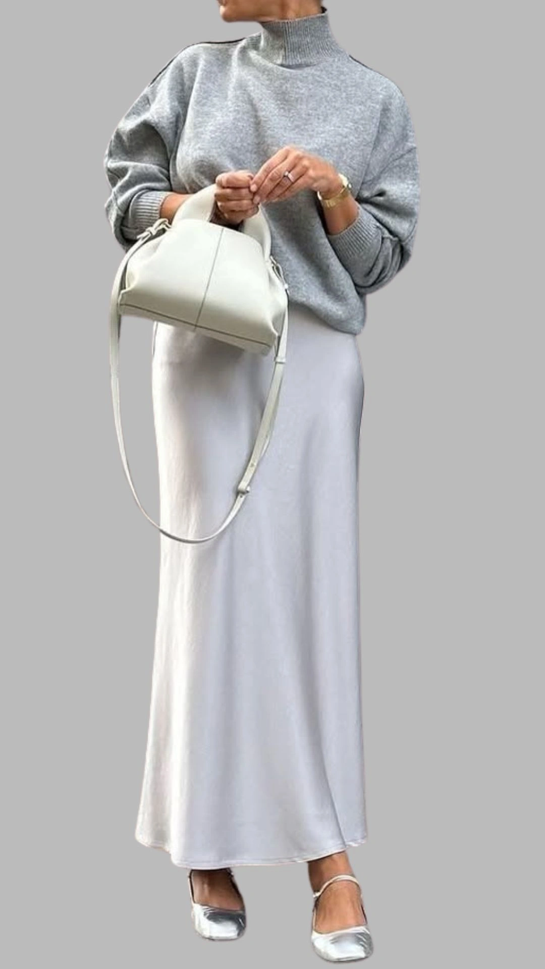 The outfit features a light grey, cropped sweater with a high neckline and relaxed, oversized sleeves paired with a floor-length, silvery-grey skirt that has a smooth finish and a straight hemline. Notable details include the cozy texture of the sweater and the elegant drape of the skirt, creating a chic monochromatic look. Accessories include a small, structured cream handbag and silver flats, enhancing the sleek aesthetic.