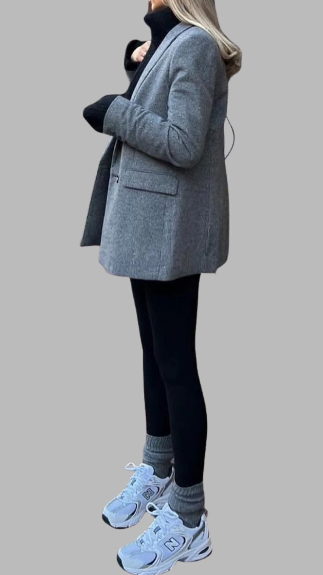 The outfit features a structured grey blazer with a notched lapel and front pockets, layered over a black turtleneck. Paired with sleek black leggings and light grey, white-trimmed sneakers, this look combines sophistication with casual comfort, perfect for versatile styling. The overall palette of grey and black is enhanced by the soft textures, creating a contemporary, chic ensemble.