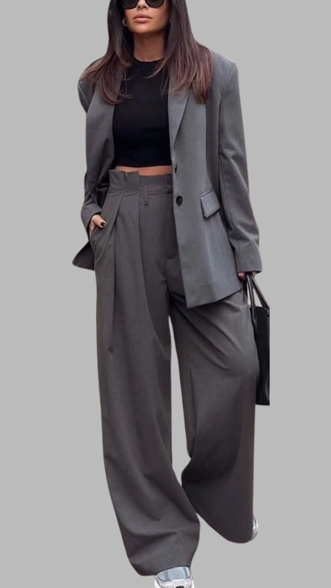 The outfit features a tailored grey blazer paired with high-waisted, wide-leg trousers, creating a sophisticated and stylish silhouette. The blazer has a notched lapel and a single button closure, while the trousers include subtle pleats and pockets for added functionality. This ensemble is complemented by a fitted black crop top, resulting in a chic, monochromatic look that balances elegance with modernity.
