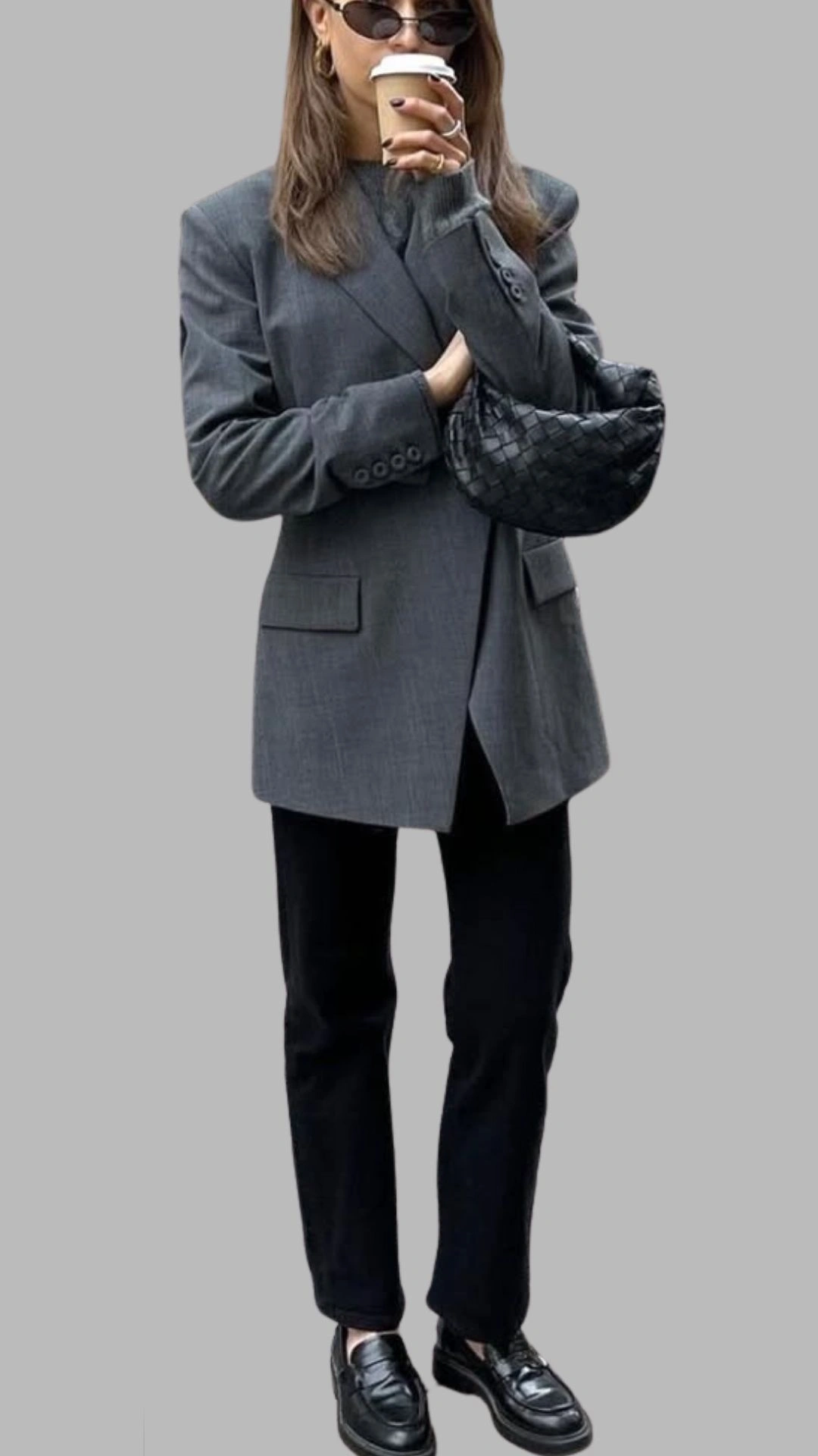 The outfit features an oversized, tailored grey blazer with a structured silhouette, complete with two front pockets and a single-breasted design. It is paired with sleek black trousers that have a straight-leg cut, creating a polished look. The ensemble is accessorized with a black woven handbag and classic black loafers for a sophisticated finish.
