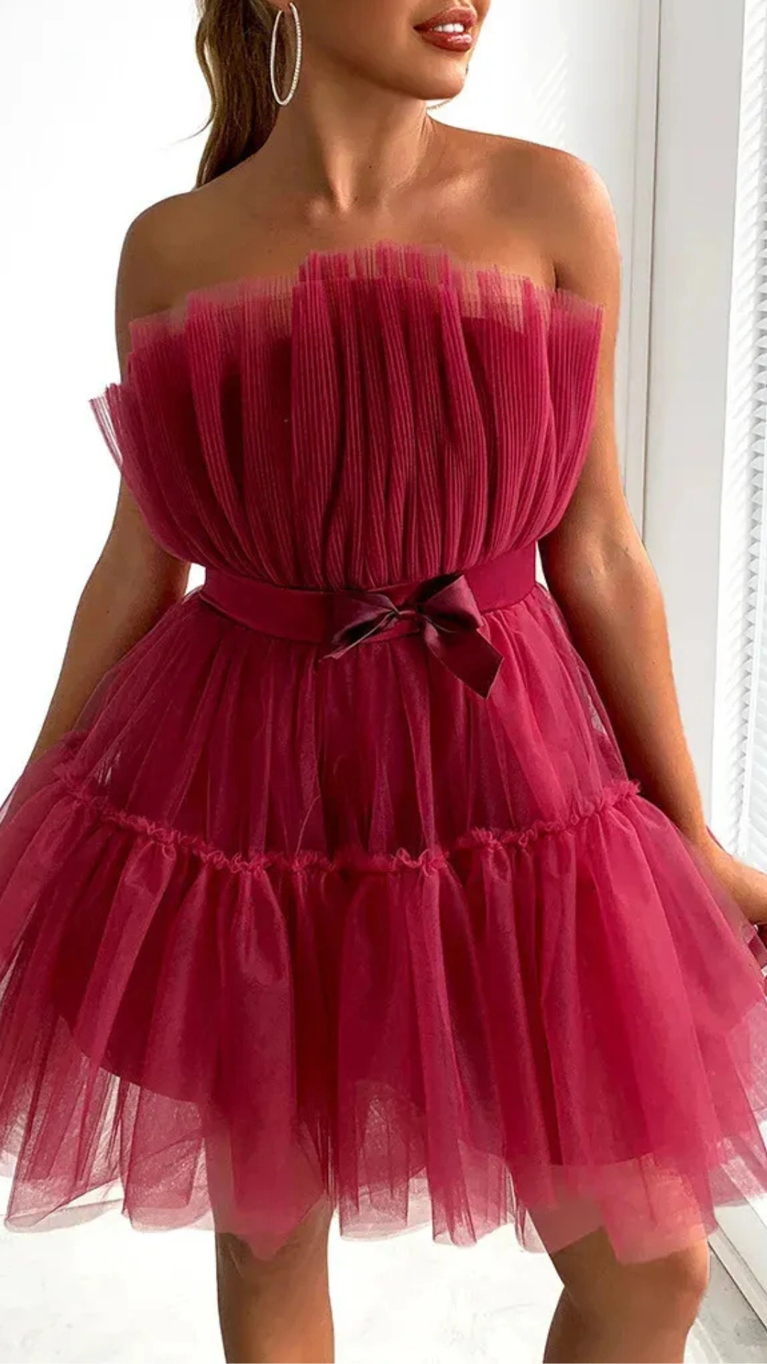 This red homecoming dress features a playful, strapless design with a fitted bodice made of pleated fabric, leading into a voluminous tulle skirt adorned with layered ruffles. Its vibrant berry hue and soft texture create a romantic and whimsical vibe, making it ideal for a festive school dance or party for teenagers.
