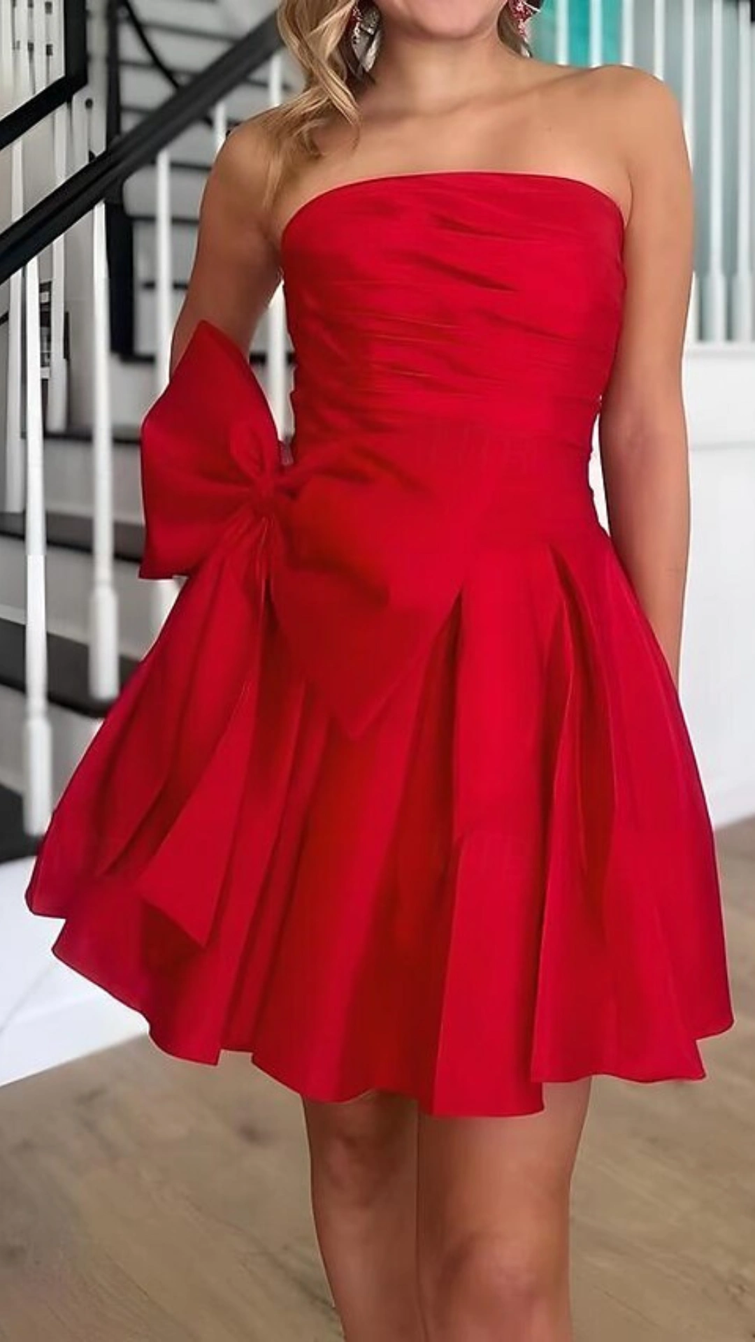 This vibrant, strapless red homecoming dress features a flattering fitted bodice with subtle ruching, leading to a playful voluminous skirt that adds a fun flair. Made of soft yet structured fabric, it is adorned with a large bow detail at the side, creating a bold, youthful vibe perfect for festive occasions like school dances or parties, aimed at teenage girls.